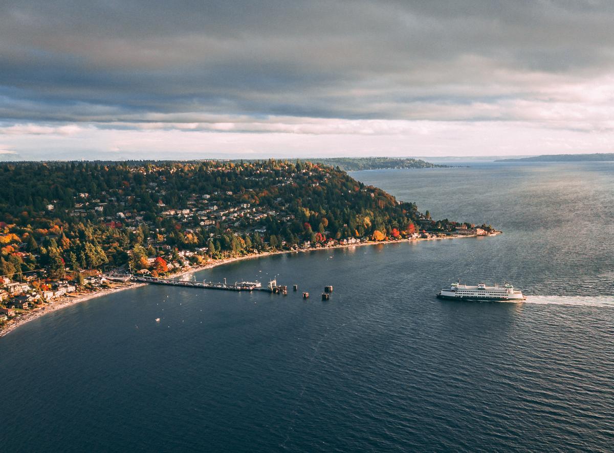 Aerial view of a ferry crossing a bay toward a small dock, with a forested coastline, houses nestled among trees, and cloudy skies above.