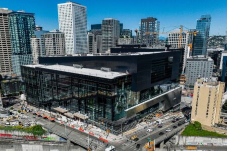 An aerial view reveals a modern, glass-fronted urban building under construction in the bustling cityscape of Seattle. Surrounded by cranes and skyscrapers, with traffic weaving below and clear blue skies above, it's a scene that captures the vibrant essence of holidays in Seattle.