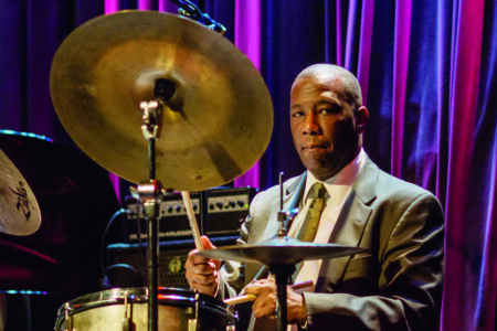 A drummer in a suit plays a drum set, focused on the performance. Dark purple curtains hang in the background, with stage lighting highlighting the musician and cymbals.