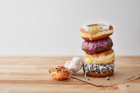A stack of assorted donuts on a wooden surface. The pile includes a powdered donut, a blueberry glazed donut, a custard-filled donut, and a coconut-topped donut. A heart-shaped donut lies beside the stack.