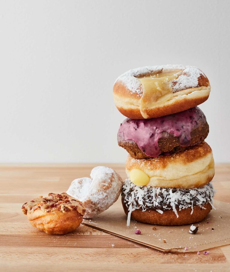 A stack of assorted donuts on a wooden surface. The top donut is glazed with powdered sugar, followed by a berry-glazed donut, a custard-filled donut, and a coconut-covered donut. Two more donuts, one heart-shaped, are placed nearby.
