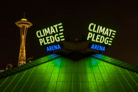 Night view of the Climate Pledge Arena with its green and blue illuminated sign, reflecting on the roof. The iconic Space Needle is visible in the background against the dark sky.