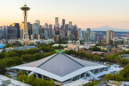 Aerial view of the Seattle skyline with the Space Needle prominently in the foreground. The Climate Pledge Arena sits surrounded by lush green trees, and Mount Rainier is visible in the distant background under a clear sky.
