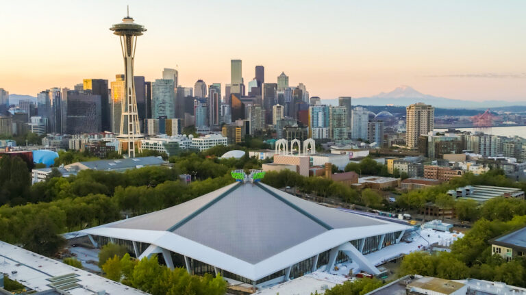 Aerial view of the Seattle skyline with the Space Needle prominently in the foreground. The Climate Pledge Arena sits surrounded by lush green trees, and Mount Rainier is visible in the distant background under a clear sky.