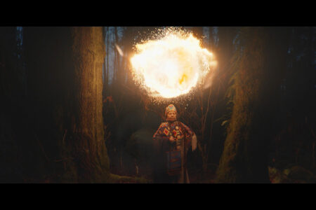A person in a forest, wearing a colorful garment, stands between two trees. They wield a large, glowing orb of fire above them, casting light in the dim, wooded surroundings.