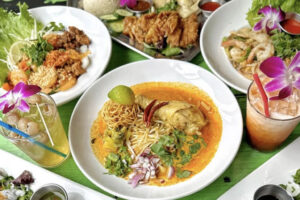 A table with colorful Thai dishes, including a bowl of curry noodles with chicken, fresh herbs, sliced red onions, lime, fried noodles on top, and drinks garnished with orchids, surrounded by fried foods and shrimp salad.