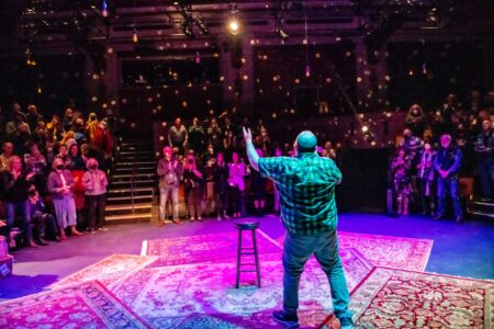 A performer stands on a stage with a spotlight, facing an audience in a theater. The audience is applauding. The stage is decorated with carpets, and hanging lights are visible in the dimly lit background.