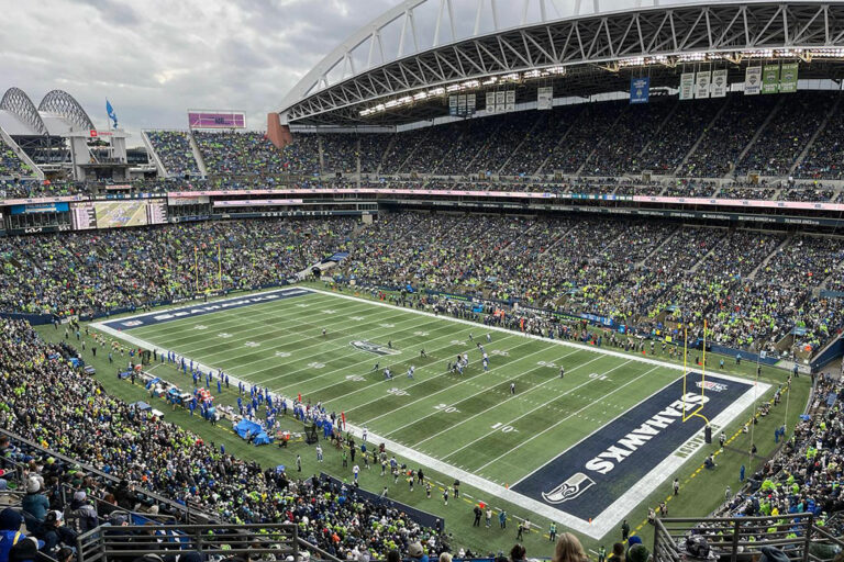 Aerial view of a crowded football stadium during a Seattle sports event. The field is lined with players, and the stands are filled with spectators wearing green and blue. The roof structure is visible, and the sky is overcast.