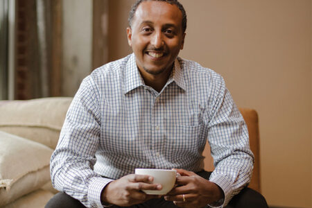 A portrait photo of Efrem Fesaha, owner of Boon Boona coffee. Efrem wears a white and blue checkered shirt and holds a white cup of coffee.