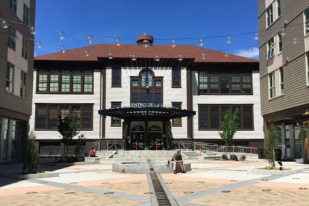 A historic building with a brown roof and white facade stands at the end of a modern courtyard. The courtyard is adorned with string lights and has two people sitting on benches. Small trees and geometric patterns decorate the plaza.