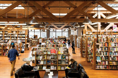 A spacious bookstore with wooden beams and floors. Shelves full of books fill the area, with people browsing and sitting on chairs reading. Large windows allow natural light to illuminate the interior.