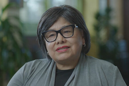 A person with glasses and short hair wearing a gray jacket and hoop earrings, smiles while sitting indoors. The background is softly blurred with green tones and natural light.