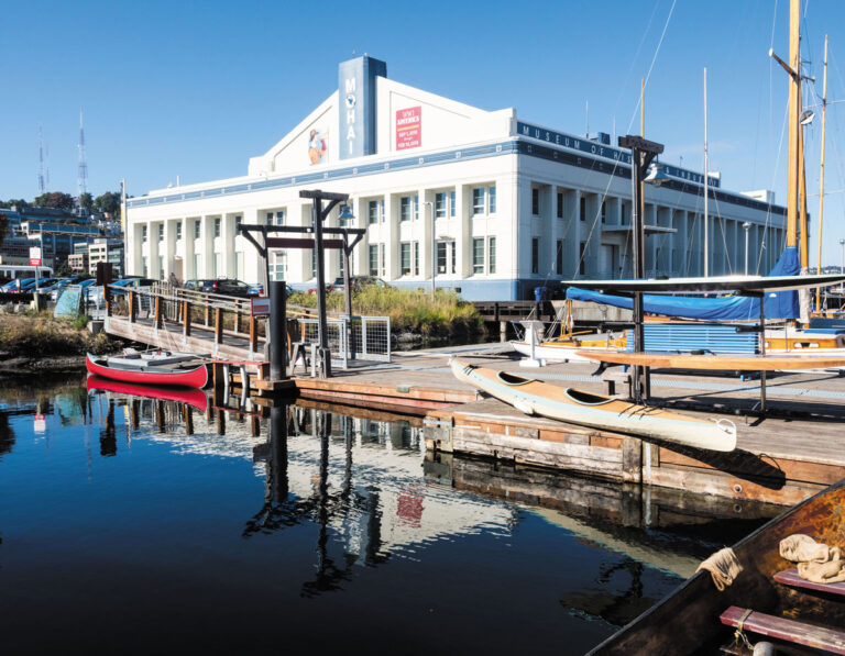 A dock with canoes and boats in the foreground, calm water reflecting the scene, and the "MOHAI" building in the background under a clear blue sky capture a serene moment in Seattle’s South Lake Union neighborhood.