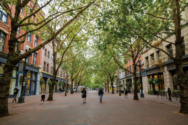 A tree-lined street with brick pavement in the Pioneer Square neighborhood, bordered by historic buildings. Several people are walking or standing along the street, and the leafy trees create a canopy overhead.