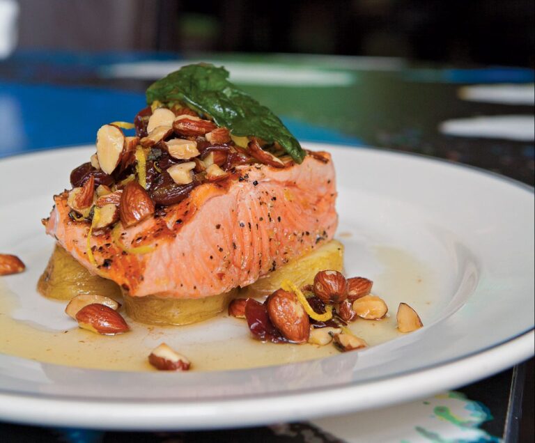 A dish featuring a grilled salmon filet topped with chopped nuts and green herbs, served over slices of cooked potatoes. The plate is garnished with a light sauce and set on a table.