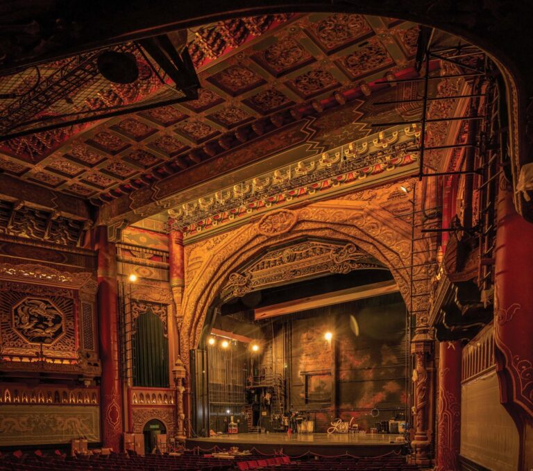 An ornate, vintage theater interior featuring intricate carvings and patterns. Rows of red seats lead to a grand proscenium stage with golden detailing. The setting is warmly lit, highlighting the richly decorated arches and ceiling.