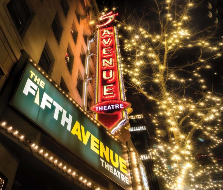 Night scene of the Fifth Avenue Theatre with a brightly lit marquee and sign. The building is adorned with festive lights, and a nearby tree is illuminated with twinkling lights, creating a warm, inviting atmosphere.