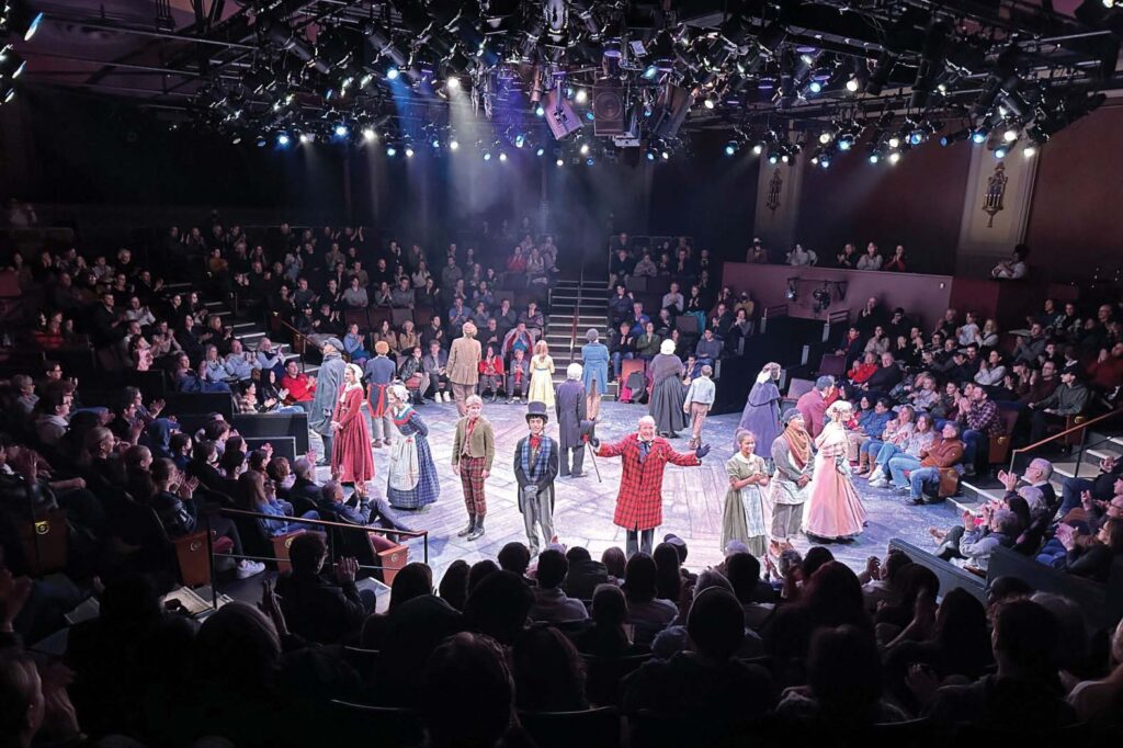 Performers in period costumes stand on a round stage surrounded by an audience. The theater is filled with spectators watching the lively, well-lit production.