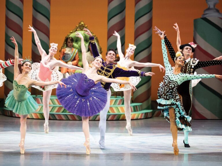 Ballet dancers in colorful costumes perform on stage, striking elegant poses. The set design is festive with candy-themed decorations in the background, adding a whimsical touch to the performance.