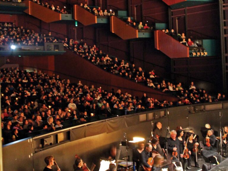 A large theater with multiple levels filled with seated audience members. An orchestra is playing in the pit, lit softly. The interior features striking geometric architecture with deep red and purple accents.