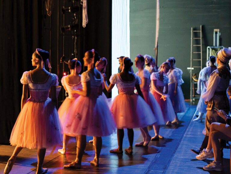 Ballet dancers in pastel tutus gather backstage, illuminated by stage lights. They are poised and preparing to perform, with a focus on the entrance. The atmosphere is tense yet graceful.