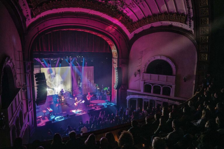 A vibrant concert scene in a historic theater with ornate architecture. The stage is illuminated with colorful lights and visuals, featuring a band performing for an engaged audience. The upper balcony and detailed ceiling are also visible.
