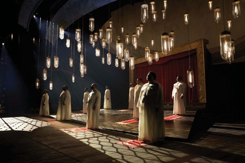 A group of people in white robes stand solemnly on a dark stage. Large hanging lights illuminate the scene, casting shadows on the wooden floor. A red curtain hangs in the background, framed by ornate gold trim.