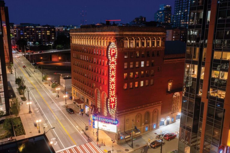 A nighttime view of a city street featuring a historic theater with a brightly lit marquee that reads "PARAMOUNT." The surrounding area is illuminated by streetlights, and modern buildings are visible in the background.