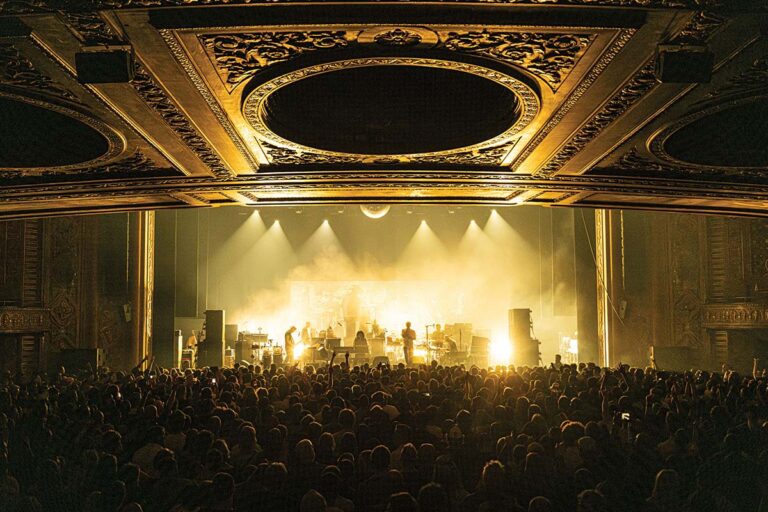 A large crowd watches a band performing on stage, illuminated by dramatic golden lighting. The ornate theater ceiling with intricate patterns adds grandeur to the scene.