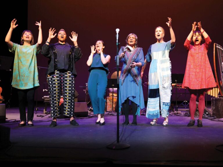 A group of six women on stage stand in a line, raising their hands in unison, with a microphone in front. They are dressed in colorful, diverse outfits, with musical instruments visible in the background. The lighting casts a warm glow on the performers.
