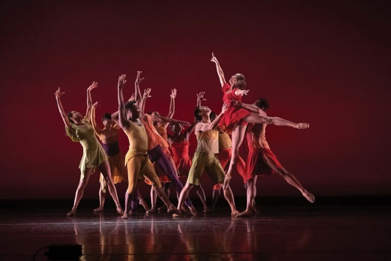 Dancers in colorful costumes perform on stage, showcasing dynamic movements and expressive poses. One dancer in a red dress is lifted gracefully above the others. The background is a rich shade of red, enhancing the vibrant scene.