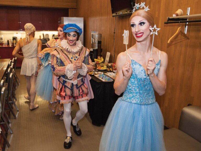 A group of performers in a backstage area, dressed in vibrant and whimsical costumes. One person in a blue dress holds a star wand, smiling. The room has a table with snacks and chairs, with costumes hanging on a rack.