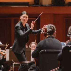 A conductor leads an orchestra on stage in a warm-lit concert hall. Musicians, dressed in formal black attire, play string instruments. The stage backdrop features softly illuminated panels and overhead lights.