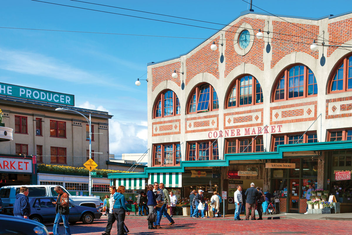 People walk and shop outside the historic Corner Market building at Pike Place Market in Seattle. The street is lively with vendors, cars, and a blue sky overhead.