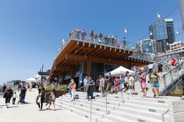 People walk and gather on a sunny day at a waterfront building with an upper deck, outdoor stairs, string lights, city buildings in the background, and tents set up along the walkway.