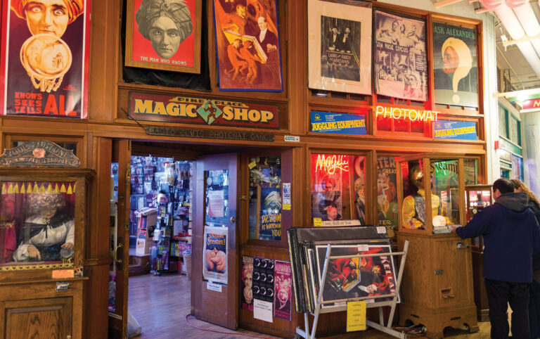 A colorful, cluttered magic shop with vintage posters, neon signs, and magic tricks on display. Two people stand near a counter, and shelves are filled with various magical items and memorabilia.