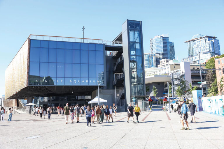 People walk outside the modern glass building of the Seattle Aquarium on a sunny day, with city skyscrapers visible in the background.