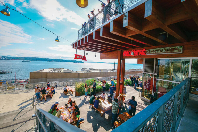 People sit at outdoor tables on a sunny patio overlooking the water, with a large “BEER” sign and brewery name visible. Some people are also on an upper balcony, enjoying the view and the lively atmosphere.