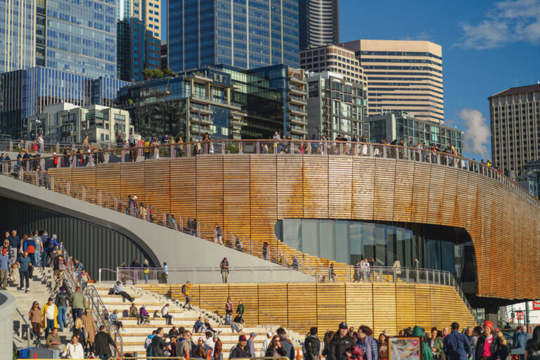 A large crowd walks and gathers on a modern wooden waterfront structure with steps, set against a backdrop of tall city skyscrapers and a blue sky.