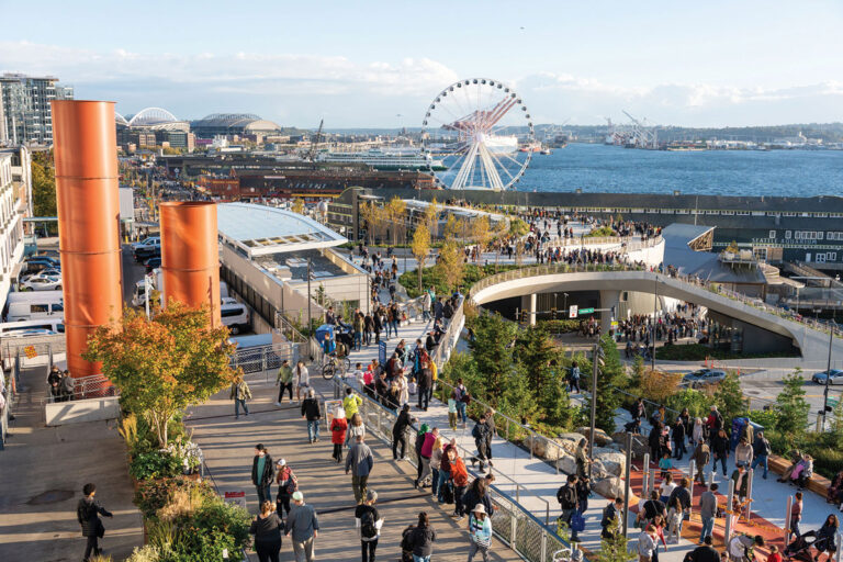 A crowded urban park with walking paths, greenery, and orange columns overlooks a waterfront with a large Ferris wheel, stadiums, and piers. People are enjoying the outdoor space on a sunny day.