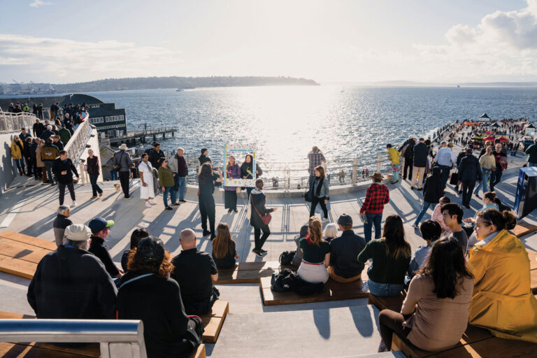 People gather on tiered seating overlooking a waterfront with bright sunlight reflecting on the water. Some are walking, others are sitting and talking, while boats and distant land are visible in the background.