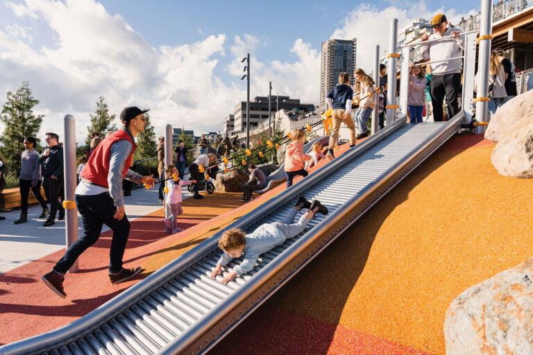 Children and adults enjoy a lively playground with a large roller slide, people climbing and sliding, and a cityscape with tall buildings in the background on a sunny day.