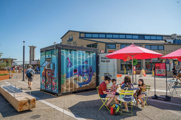 A group of people sit at a table under red umbrellas near a colorful painted container in an outdoor plaza, with a large building and other visitors in the background on a sunny day.