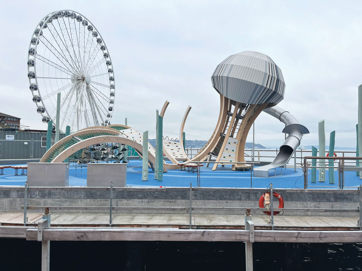 A modern waterfront playground with abstract climbing structures and a large dome slide, set near a Ferris wheel and water, under a cloudy sky. Safety railings and a life preserver are visible in the foreground.
