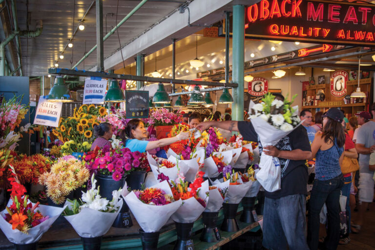 A bustling flower market with vibrant bouquets and buckets of colorful flowers. A vendor hands a bouquet to a customer as other shoppers browse. Signs and lights hang above the busy stall.