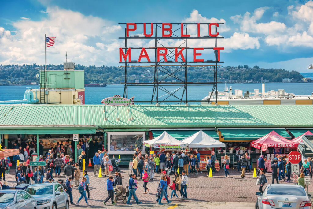 A bustling crowd walks in front of Seattle’s Pike Place Market, with the iconic red “Public Market” sign above green rooftops, vendor tents, and views of water and trees in the background under a partly cloudy sky.
