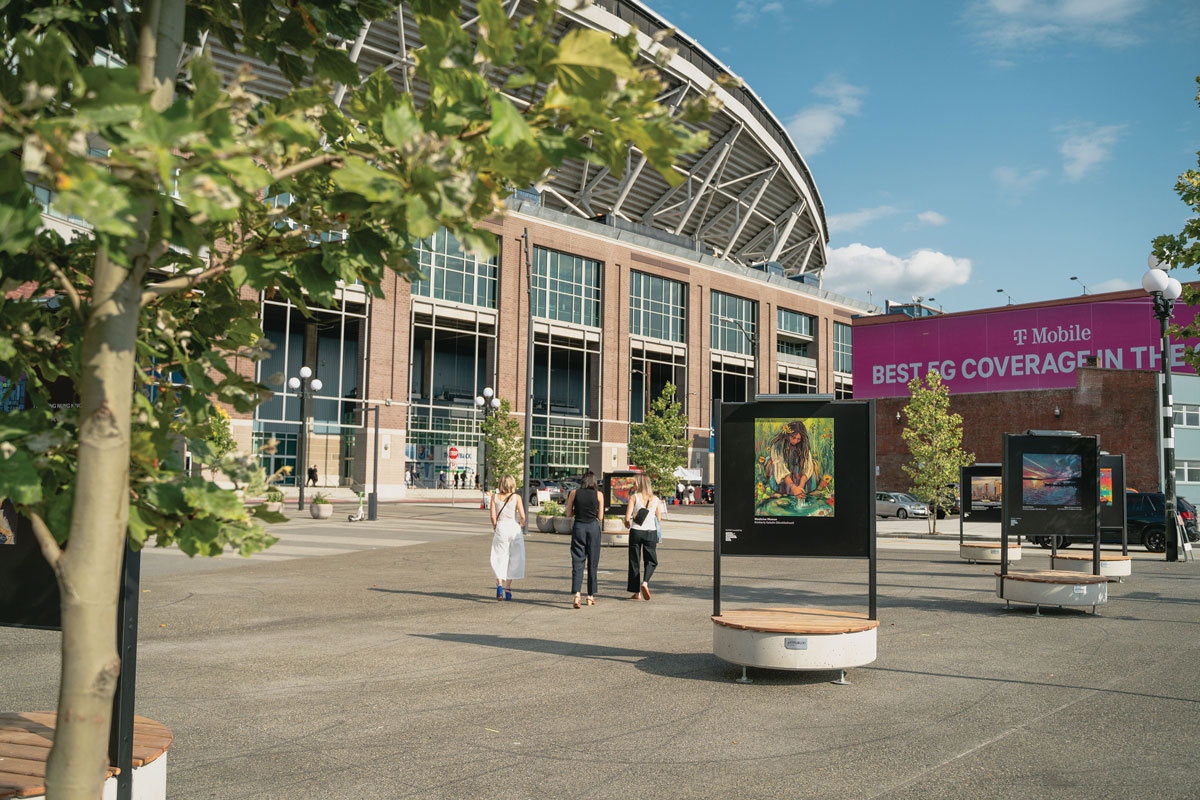 Three people walk towards an outdoor art exhibit displayed on black stands near a stadium, with trees in the foreground and a large "Best 5G Coverage in the..." T-Mobile sign in the background.