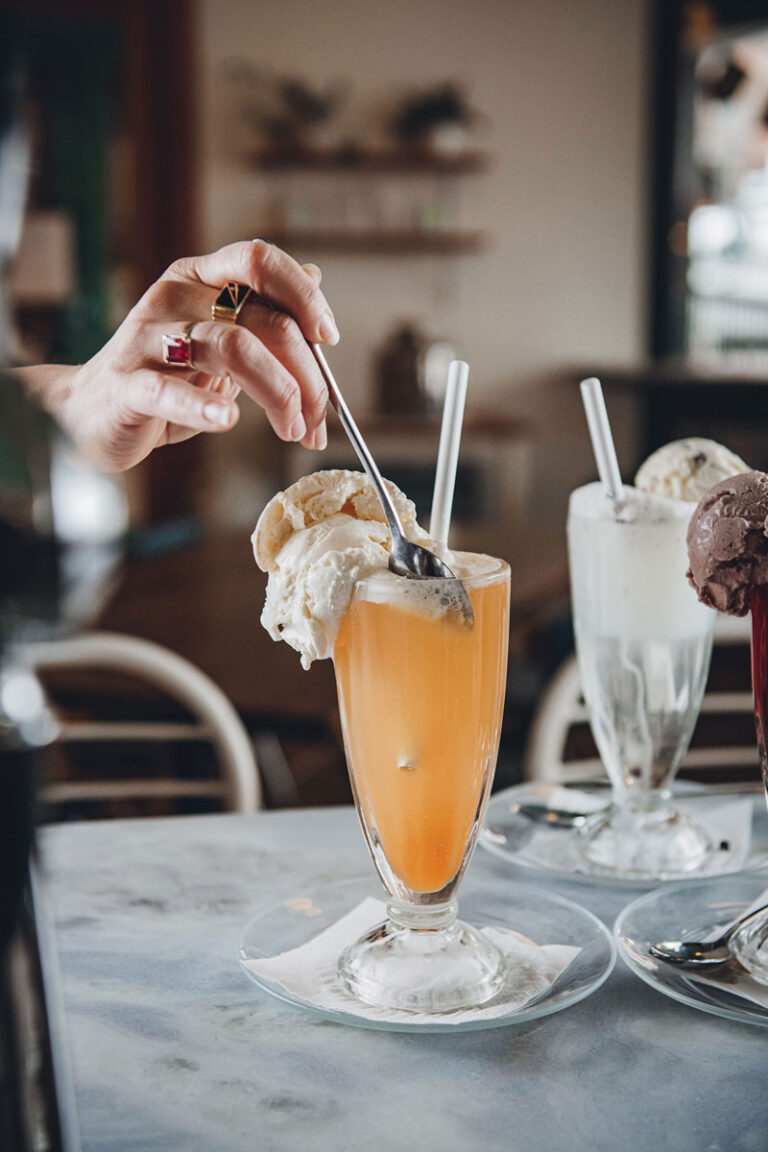 A hand holding a spoon scoops ice cream from a tall glass of orange soda float, with two other ice cream floats in the background on a marble table.