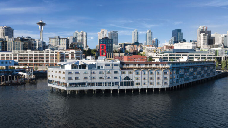 A large waterfront building with a red "B" sign sits on the water in front of a city skyline, with the Space Needle visible in the background under a clear blue sky.