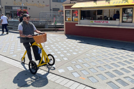 A person using a yellow mobility aid sits on the sidewalk in front of a red building with yellow awning.